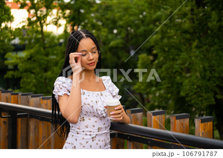 Portrait of a young attractive African woman with glasses holding a paper cup of coffee and looking at it on the street during the day in the park in summer Portrait of a young attractive African woman with glasses holding a paper cup of coffee and looking at it on the street during the day in the park in summer 106791787