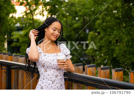 Portrait of a young attractive African woman with glasses holding a paper cup of coffee and looking at it on the street during the day in the park in summer Portrait of a young attractive African woman with glasses holding a paper cup of coffee and looking at it on the street during the day in the park in summer 106791790