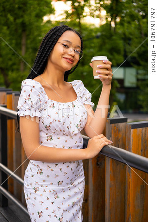 African young attractive woman in glasses stands on a bridge with a paper cup of coffee on the street during the day in the park in summer and looks at the camera African young attractive woman in glasses stands on a bridge with a paper cup of coffee on the street during the day in the park in summer and looks at the camera 106791797