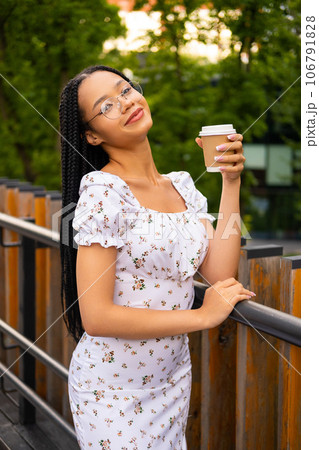 African young attractive woman in glasses stands on a bridge with a paper cup of coffee on the street during the day in the park in summer and looks at the camera African young attractive woman in glasses stands on a bridge with a paper cup of coffee on the street during the day in the park in summer and looks at the camera 106791828