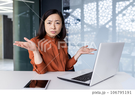 Portrait of a young Asian woman working in the office on a laptop and worriedly spreading her hands to the camera. Problems with work, study. 106795191