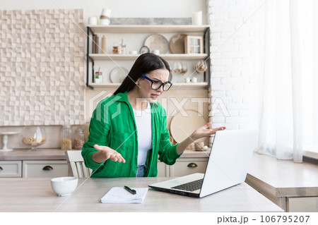 Worried and upset young woman sitting in the kitchen at home and talking on the phone on a laptop, reading a message and email, throwing up her hands in frustration. 106795207
