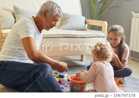 Grandfather and granddaughters playing with toys sitting on floor near sofa in living room spending happy timew together enjoying games at home interior. Grandfather and granddaughters playing with toys sitting on floor near sofa in living room spending happy timew together enjoying games at home interior. 106797370