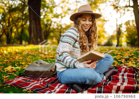 Beautiful curly woman in hat on mat with book in autumn park. Relaxation, solitude with nature. Beautiful curly woman in hat on mat with book in autumn park. Relaxation, solitude with nature. 106798968