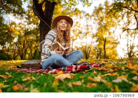 Beautiful curly woman in hat on mat with book in autumn park. Relaxation, solitude with nature. 106798969