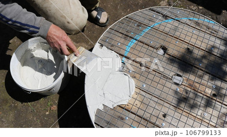the master applies an elastic solution with a narrow spatula to a round mesh surface, creating unique furniture from various materials, applying mastic with a knife to a surface with a metal grid the master applies an elastic solution with a narrow spatula to a round mesh surface, creating unique furniture from various materials, applying mastic with a knife to a surface with a metal grid 106799133