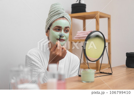 Positive woman applying green clay mask on nose with brush looking in table mirror young lady in terry bathrobe doing face cleansing procedure at home Positive woman applying green clay mask on nose with brush looking in table mirror young lady in terry bathrobe doing face cleansing procedure at home 106799410