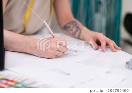 Woman draws dress model sitting at desk in clothes atelier talented designer makes sketch of stylish apparel for women in fashion studio closeup sewing industry Woman draws dress model sitting at desk in clothes atelier talented designer makes sketch of stylish apparel for women in fashion studio closeup sewing industry 106799460