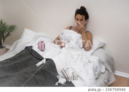 An ailing woman sits in bed, wiping her nose and holding medical drops to combat her illness.  106799986