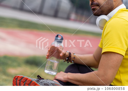 Athlete in colorful t-shirt, with headphones on, looks downhearted at his water bottle during a training break at the sports arena. Athlete in colorful t-shirt, with headphones on, looks downhearted at his water bottle during a training break at the sports arena. 106800858