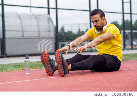 Energetic athlete, with earphones, stretches in butterfly position on stadium track in a lush city park's sports arena. 106800860