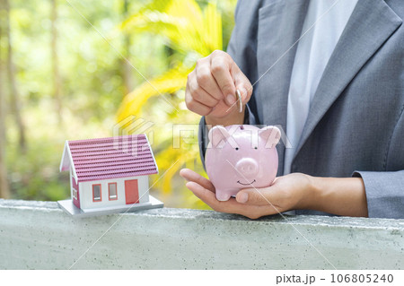 Businesswoman hands putting coin into piggybank, the home model stands in front of home background, Saving money for buy real Estate concept Businesswoman hands putting coin into piggybank, the home model stands in front of home background, Saving money for buy real Estate concept 106805240