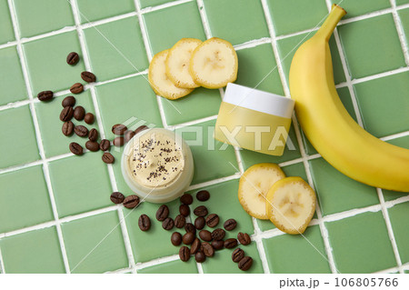 A yellow cream jar decorated with fresh banana, banana slices and brown coffee beans on green ceramic background. Mockup scene for cosmetic skin care, face mask and hair mask A yellow cream jar decorated with fresh banana, banana slices and brown coffee beans on green ceramic background. Mockup scene for cosmetic skin care, face mask and hair mask 106805766