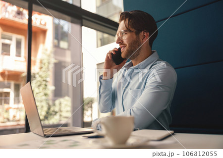 Male boss talking phone with client while sitting in modern office and working on laptop 106812055