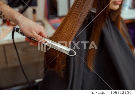 Closeup of hairdresser hand holding hair straightener and making hairstyle for female client 106812156