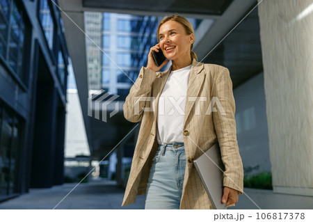 Smiling business woman holding laptop and talking phone standing on modern building background Smiling business woman holding laptop and talking phone standing on modern building background 106817378