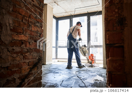 Man finishing concrete surface with floor screed grinder machine in room with large window. Back view of male worker using troweling machine while screeding floor in apartment under renovation. 106817414