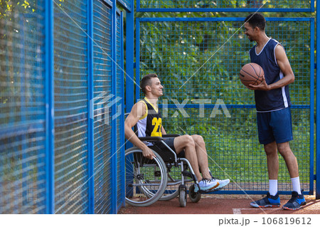 Man playing basketball with disabled friend in wheelchair at outdoor court. 106819612