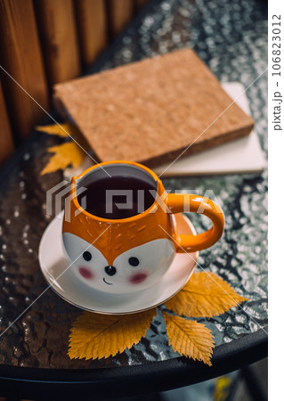 Autumn cozy still life on balcony, terrace. Fall cozy set with autumn fall leaves, cup of coffee or tea on brown books and notepads on balcony, terrace glass table. Relaxing still life concept. 106823012