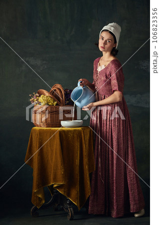 Countryside morning. Beautiful woman, medieval maid in historical attire pouring milk against vintage green background 106823356