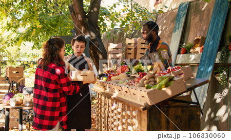 Customers buying healthy fresh bio food without pesticides, standing at farmers market counter. Smiling local vendors greeting clients and selling homegrown fruits and veggies from farm. Customers buying healthy fresh bio food without pesticides, standing at farmers market counter. Smiling local vendors greeting clients and selling homegrown fruits and veggies from farm. 106830060