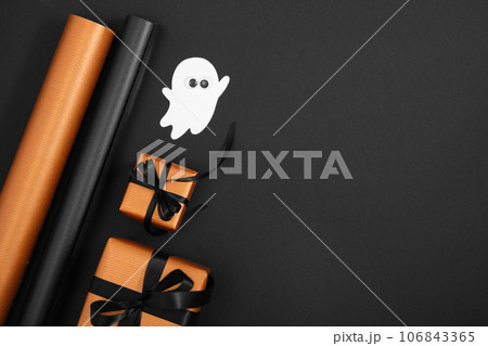 Orange gift boxes with black ribbons, rolls of wrapping paper and ghost of white cardboard on black background. Halloween gift wrapping. 106843365