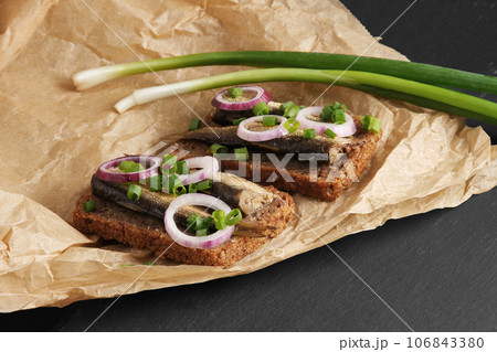 Sandwiches with sprats, red and green onions on baking paper on black stone background. Appetizer, seafood, front view Sandwiches with sprats, red and green onions on baking paper on black stone background. Appetizer, seafood, front view 106843380