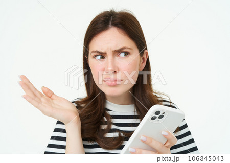 Portrait of angry, confused young woman shrugging shoulders while using mobile phone, holding smartphone with annoyed face expression, frowning, standing over white background Portrait of angry, confused young woman shrugging shoulders while using mobile phone, holding smartphone with annoyed face expression, frowning, standing over white background 106845043