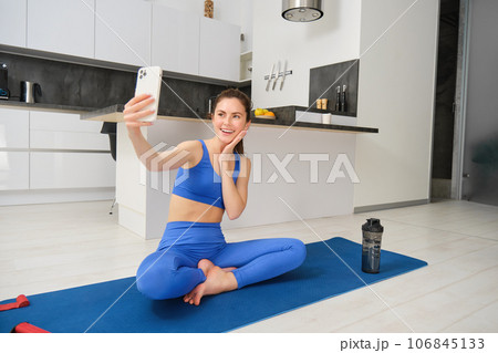 Young smiling woman, fitess girl doing workout from home, sits on yoga mat with smartphone, taking selfie, records sport vlog indoors Young smiling woman, fitess girl doing workout from home, sits on yoga mat with smartphone, taking selfie, records sport vlog indoors 106845133