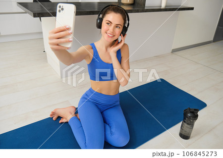 Beautiful and stylish sportswoman, posing for selfie, listens music in wireless headphones, holds smartphone, sits on fitness mat, workout from home 106845273
