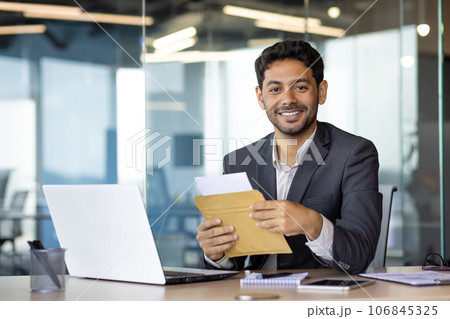 Portrait of young successful businessman at workplace, man received mail envelope notification letter, man smiling and looking at camera, satisfied with achievement results boss in business suit . Portrait of young successful businessman at workplace, man received mail envelope notification letter, man smiling and looking at camera, satisfied with achievement results boss in business suit . 106845325
