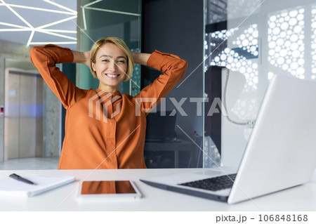 Well done woman completed work, businesswoman at workplace inside office smiling and looking at camera, female worker threw hands behind head satisfied with achievement results. 106848168