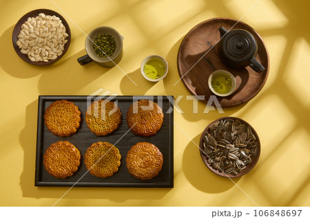 Mooncake is a traditional cake on mid autumn festival, placed on black tray displayed with dishes of pumpkin seed and sunflower seed on yellow background. Advertising photo Mooncake is a traditional cake on mid autumn festival, placed on black tray displayed with dishes of pumpkin seed and sunflower seed on yellow background. Advertising photo 106848697