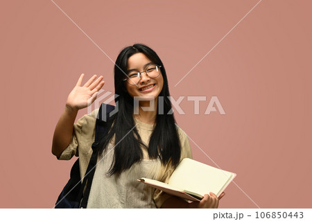 Cheerful schoolgirl with backpack holding books and waving hand at camera. Education, learning and back to school concept Cheerful schoolgirl with backpack holding books and waving hand at camera. Education, learning and back to school concept 106850443