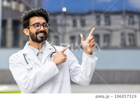 A young Hindu doctor from outside the clinic smiles and looks away, the man points his fingers in the direction, smiles with satisfaction and the doctor with a medical coat and a stethoscope. A young Hindu doctor from outside the clinic smiles and looks away, the man points his fingers in the direction, smiles with satisfaction and the doctor with a medical coat and a stethoscope. 106857329
