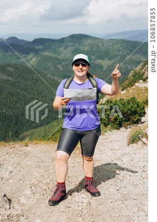 Stylish sport woman holding paper map, wearing backpack and hat looking at mountain view while relaxing in nature. Travel and wanderlust concept. Amazing chill moment. Full body 106857855