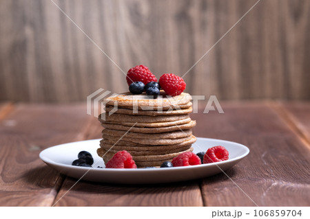 A stack of pancakes with fresh raspberries and blueberries on a light plate on a wooden background 106859704