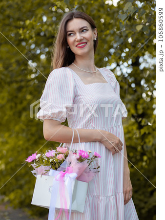 Beautiful girl with flowers, in a dress, glasses and sneakers. A beautiful bouquet of flowers in a box in the hands of a beautiful girl who walks along the street on a sunny day. Beautiful girl with flowers, in a dress, glasses and sneakers. A beautiful bouquet of flowers in a box in the hands of a beautiful girl who walks along the street on a sunny day. 106860599