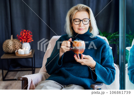 Middle age woman enjoying with aroma of hot coffee drink and relaxing in scandy style hygge interior home with fall mood decor. Lady dreaming, calm mood without stress, well being alone. Cozy autumn 106861739