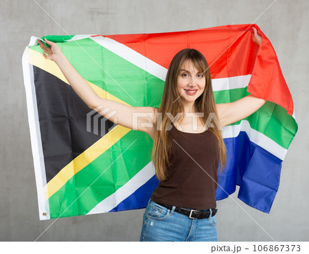 Portrait of confident smiling young female student standing against gray wall with national flag of South Africa 106867373