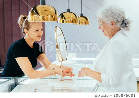 Elderly grey haired woman buying some jewelry. Woman owner, saleswoman of jewelry shop presenting for senior female consumer golden ring with diamond. 106876081