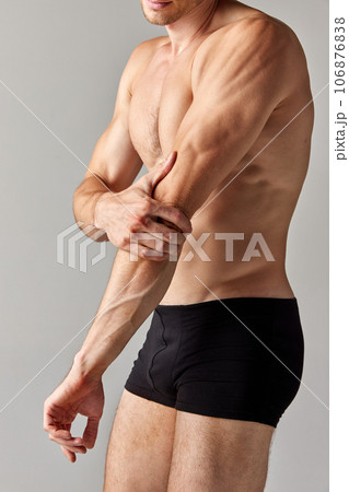 Hand pain, sprain. Cropped image of muscular man with relief body in underwear holding elbow against grey studio background Hand pain, sprain. Cropped image of muscular man with relief body in underwear holding elbow against grey studio background 106876838