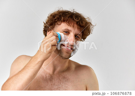 Shirtless mature man with curly hair taking care after skin, washing face with foam and brush against grey studio background 106876878