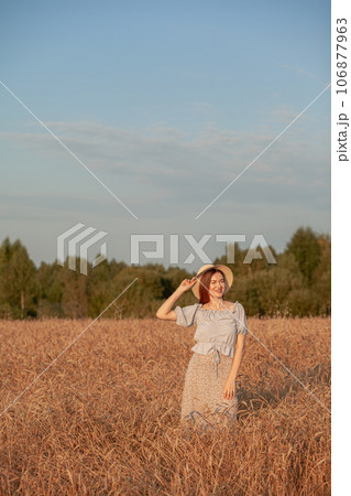 A young beautiful girl with braces on her teeth and long hair poses in a wheat field in the summer at sunset. The girl holds a hat in her hand against the background of a wheat field. 106877963