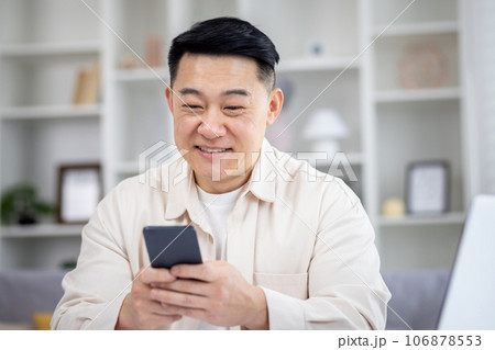 Mature Asian man sitting at table at home, man using phone, typing message on smartphone, app user and browsing internet in home cinema room, smiling contentedly. Mature Asian man sitting at table at home, man using phone, typing message on smartphone, app user and browsing internet in home cinema room, smiling contentedly. 106878553
