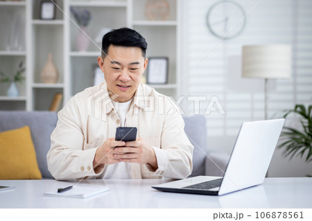 Mature Asian man sitting at table at home, man using phone, typing message on smartphone, app user and browsing internet in home cinema room, smiling contentedly. Mature Asian man sitting at table at home, man using phone, typing message on smartphone, app user and browsing internet in home cinema room, smiling contentedly. 106878561