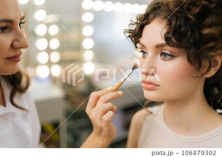 Closeup face of young sensual woman sitting at dressing table and looking in mirror while makeup artist applying concealer with cosmetic brush. Female stylist doing makeup for client in beauty salon. 106879302