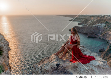 Woman sunset sea red dress, side view a happy beautiful sensual woman in a red long dress posing on a rock high above the sea on sunset. Woman sunset sea red dress, side view a happy beautiful sensual woman in a red long dress posing on a rock high above the sea on sunset. 106880560