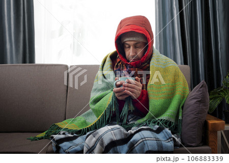 Middle age male model in hat parka and plaid hold coffee cup sitting on the sofa in his cold living room Middle age male model in hat parka and plaid hold coffee cup sitting on the sofa in his cold living room 106883339