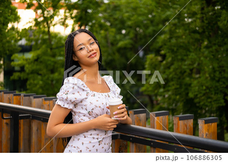 African young attractive woman in glasses stands on a bridge with a paper cup of coffee on the street during the day in the park in summer and looks at the camera 106886305